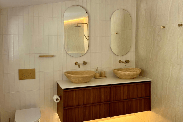 Modern bathroom with dual oval mirrors, beige vessel sinks, wooden vanity, and wall-mounted faucets.