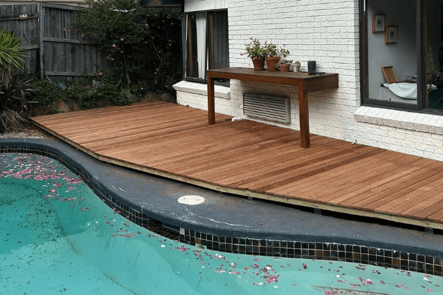Wooden deck next to a swimming pool with scattered petals and a table with potted plants by a white brick wall.