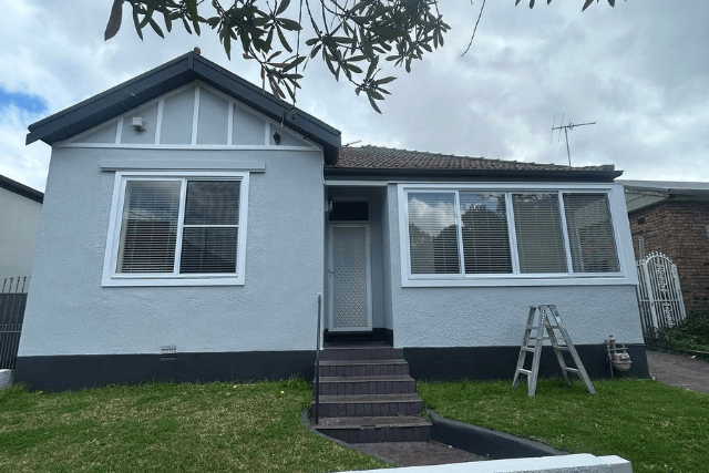 Single-story gray house with white-framed windows and a front door with steps and a ladder nearby.