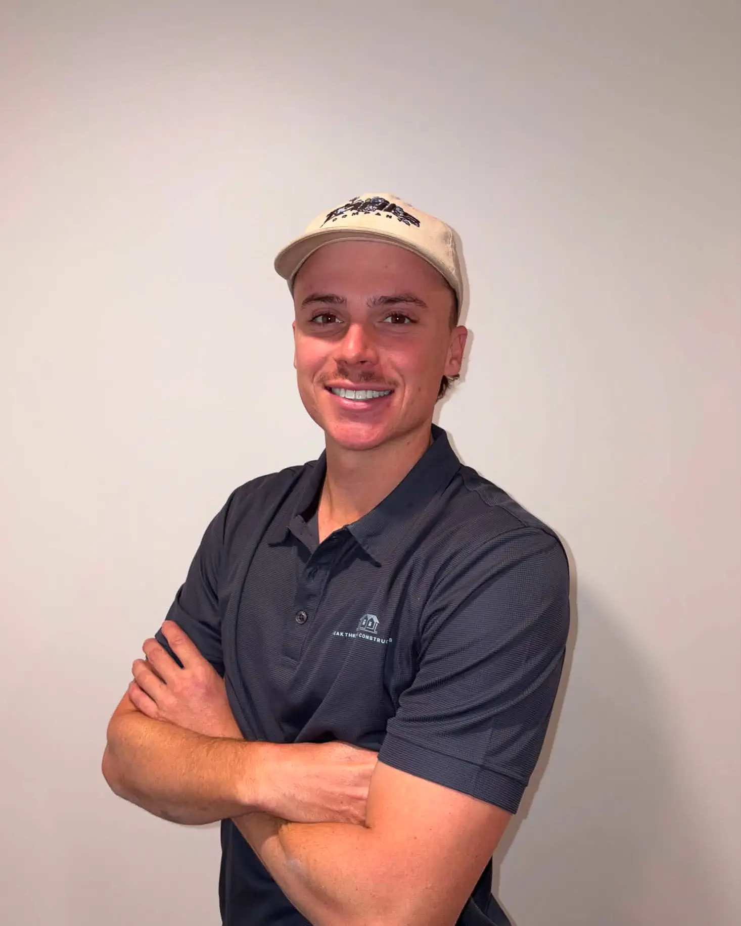 Smiling man with a mustache wearing a light cap and black polo shirt, arms crossed in front of a plain wall.
