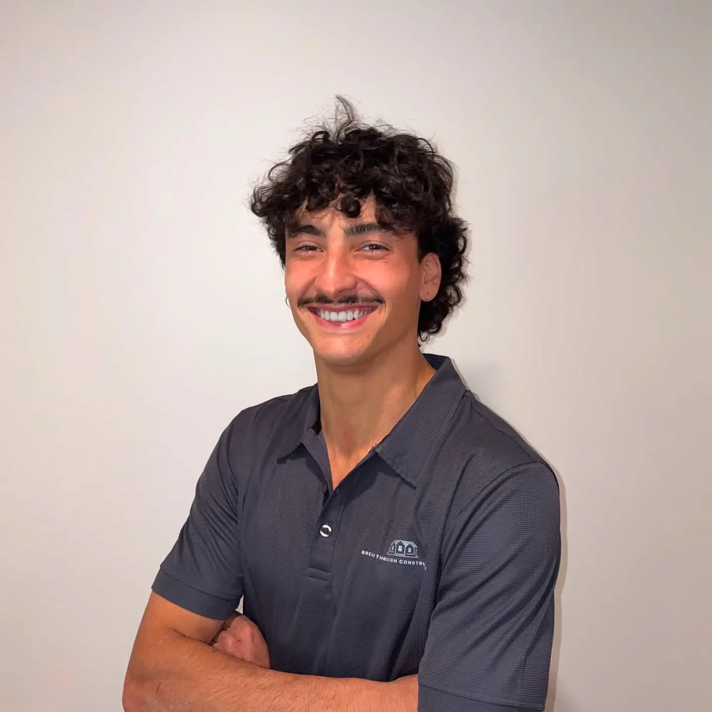 Smiling man with curly hair and mustache wearing a dark polo shirt with folded arms.