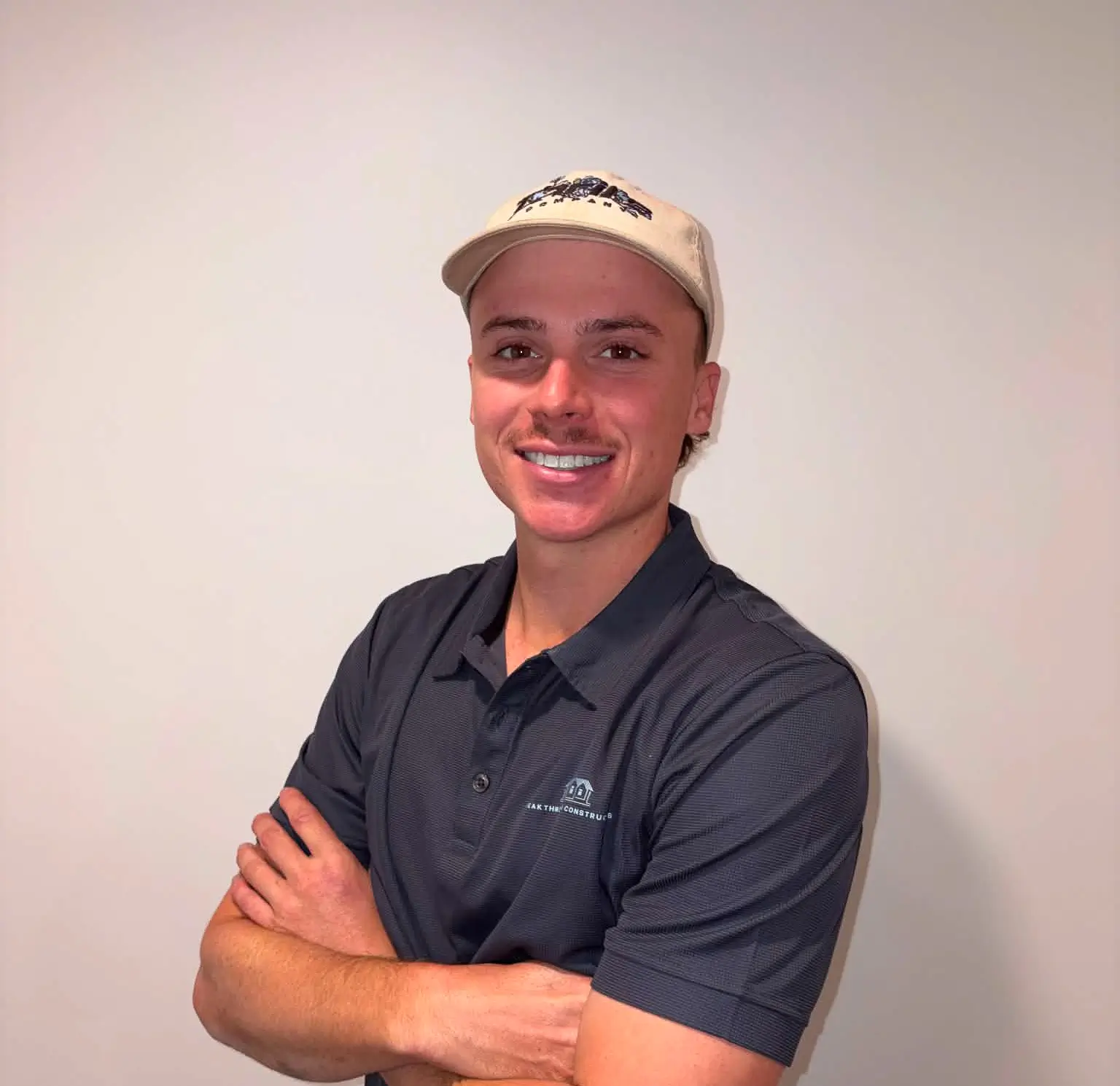 Smiling man wearing a light beige cap and navy blue shirt with crossed arms against a plain wall.