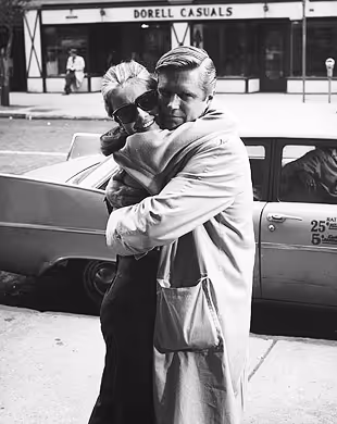 Black and white photo of a man and woman hugging on a city sidewalk near a parked car, Mr. C Miami – Coconut Grove, luxury hotel.