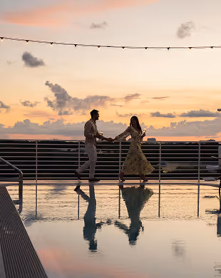 Couple holding hands and walking by a pool at sunset with string lights above them, Mr. C Miami – Coconut Grove, luxury hotel.