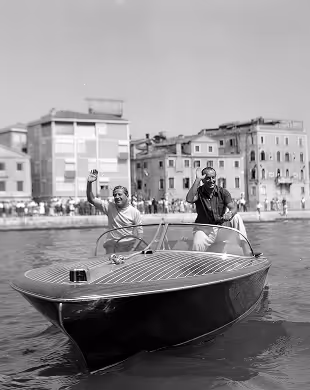 Two men waving from a classic wooden speedboat on water with buildings in the background, Mr. C Miami – Coconut Grove, luxury hotel.