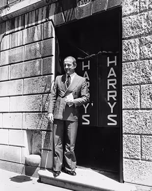 Man in suit holding papers and cane standing in front of a doorway with 'HARRY'S' signs, Mr. C Miami – Coconut Grove, luxury hotel.