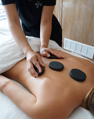 Person receiving hot stone massage with three black stones on their back and therapist's hands applying pressure, Mr. C Miami – Coconut Grove, luxury hotel.