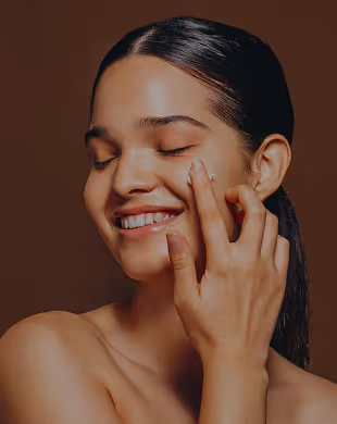 Smiling woman with eyes closed applying cream to her cheek with fingers against brown background, Mr. C Miami – Coconut Grove, luxury hotel.