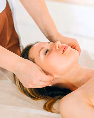 Woman relaxed with eyes closed receiving a neck and head massage on a white bed, Mr. C Miami – Coconut Grove, luxury hotel.
