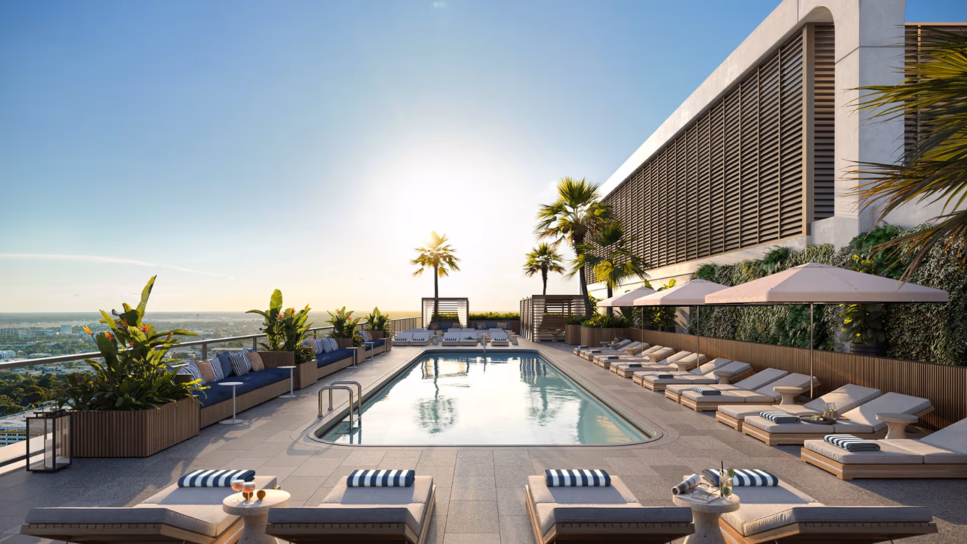 Piscina panoramica illuminata dal sole con sedie a sdraio, ombrelloni, palme e vista sulla città al tramonto.