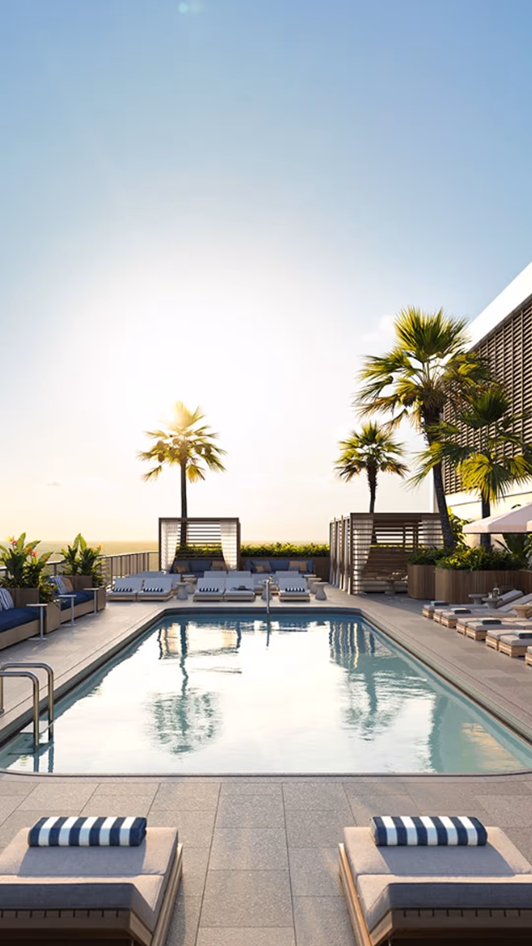 Zona piscina illuminata dal sole con sedie a sdraio, asciugamani a righe, palme e cabine in un resort moderno.