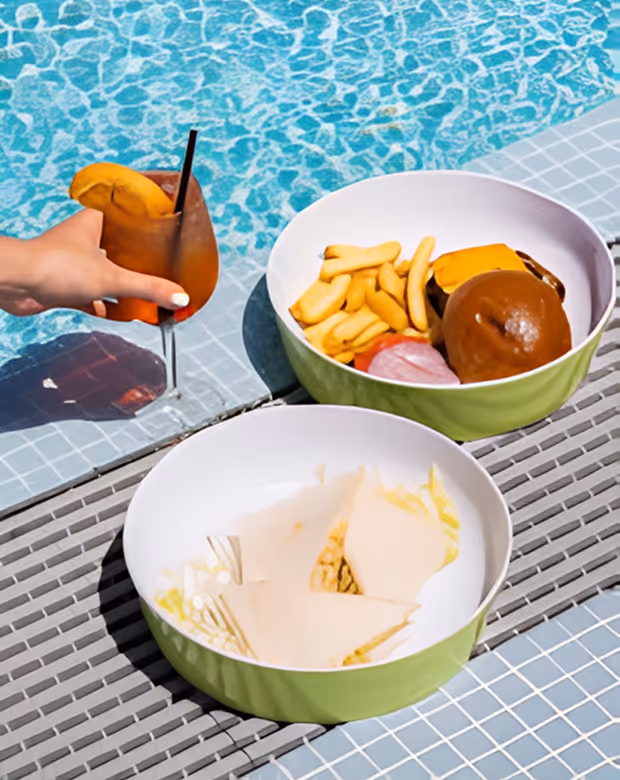 Two bowls with snacks by a pool, one with chips and dip, the other with fries and a burger, and a hand holding a drink, Mr. C Miami – Coconut Grove, luxury hotel.