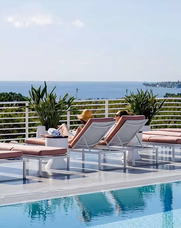 Two people relaxing on lounge chairs by a pool with ocean view and plants under clear sky, Mr. C Miami – Coconut Grove, luxury hotel.