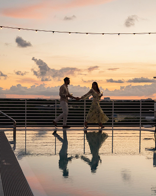Coppia che si tiene per mano e cammina vicino a una piscina sul tetto al tramonto con luci a corda sopra.