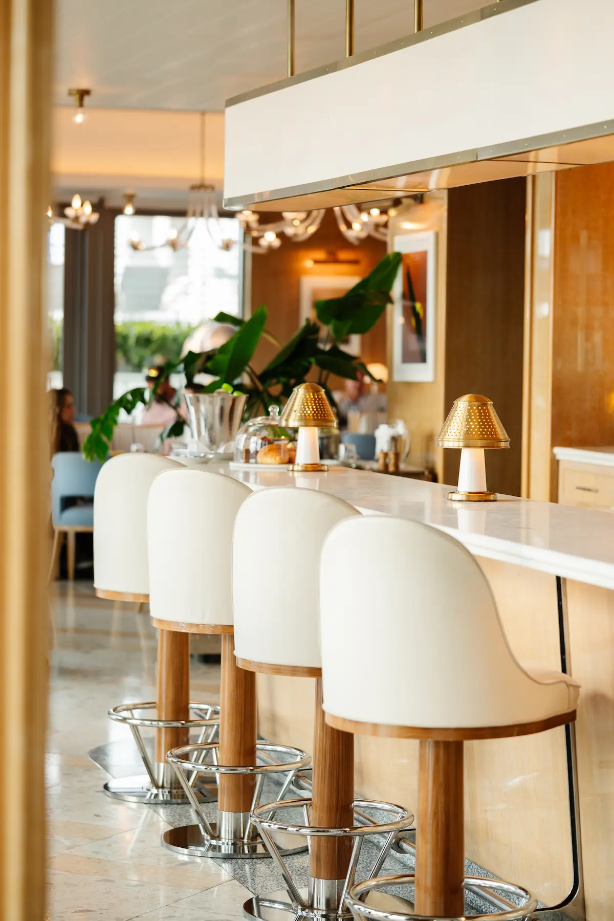 Four white cushioned bar stools with wooden legs at a marble countertop with gold lamps, Mr. C Miami – Coconut Grove, luxury hotel.