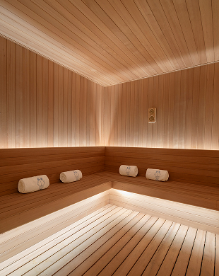 Interno di una sauna in legno con panche angolari, tre asciugamani arrotolati e calda illuminazione ambientale.