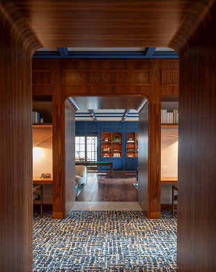 Interno rivestito in legno con arco, moquette a motivi geometrici, scrivanie e parete blu con scaffali sullo sfondo.