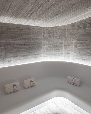 Panca curva bianca minimalista con tre asciugamani arrotolati sotto illuminazione indiretta a soffitto.