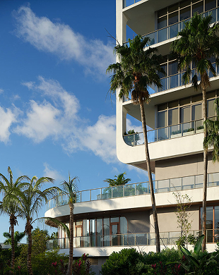 Esterno dell'edificio moderno con balconi, ringhiere in vetro, alte palme e un cielo blu con nuvole.