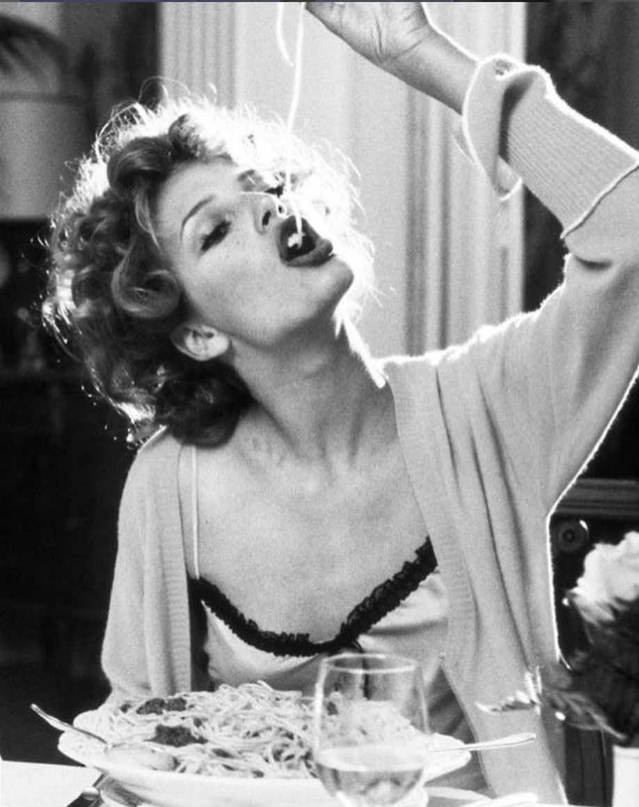 Donna con i capelli ricci che mangia spaghetti con la testa inclinata all'indietro e le tagliatelle che le penzolano dalla bocca.