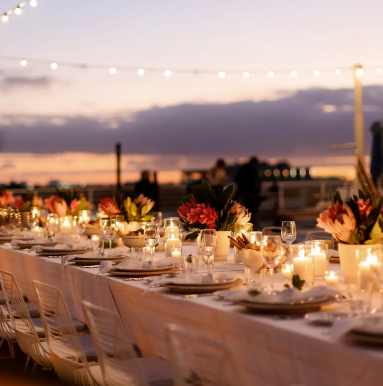 Elegante tavolo da pranzo per esterni con centrotavola floreali e candele al tramonto con luci a filo sopra.