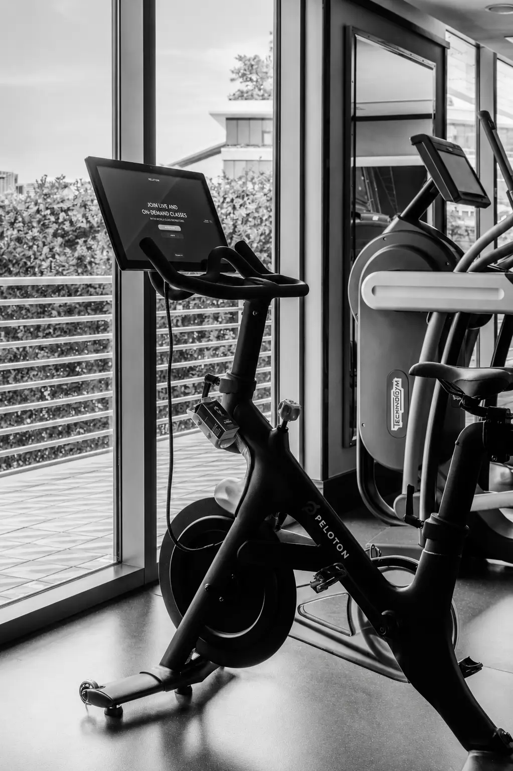 Bicicleta estática Peloton con pantalla en el gimnasio cerca de grandes ventanales que muestran vegetación al aire libre.