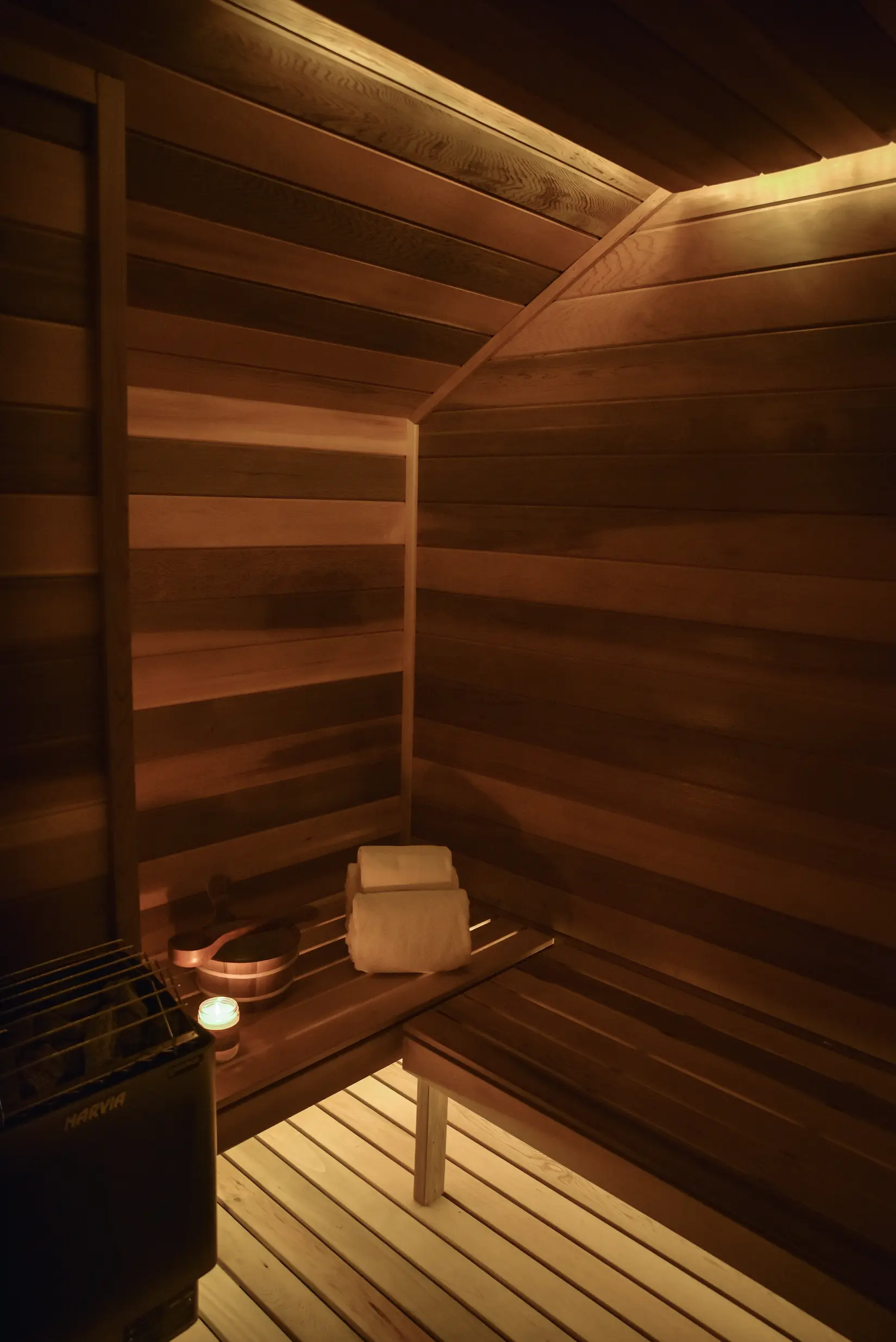 Interior de sauna con paneles de madera con bancos, toallas enrolladas, cucharón de agua, vela y calentador.
