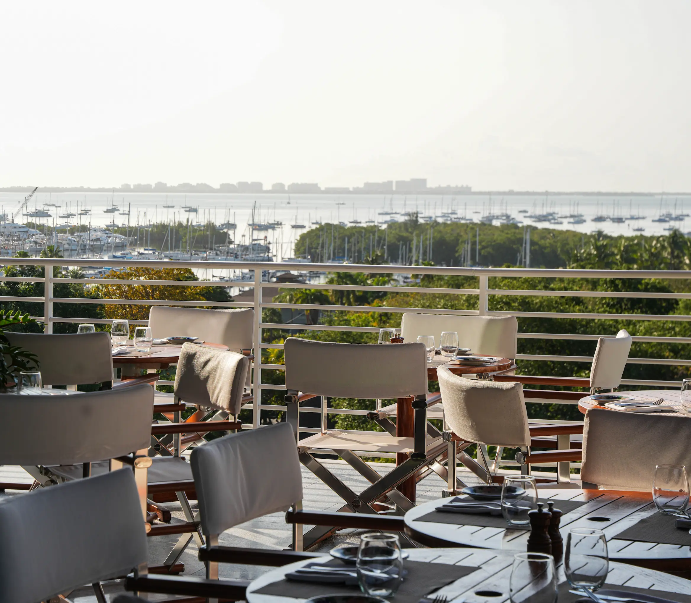 Salle à manger extérieure avec tables, chaises et verrerie donnant sur un port de plaisance avec des bateaux.