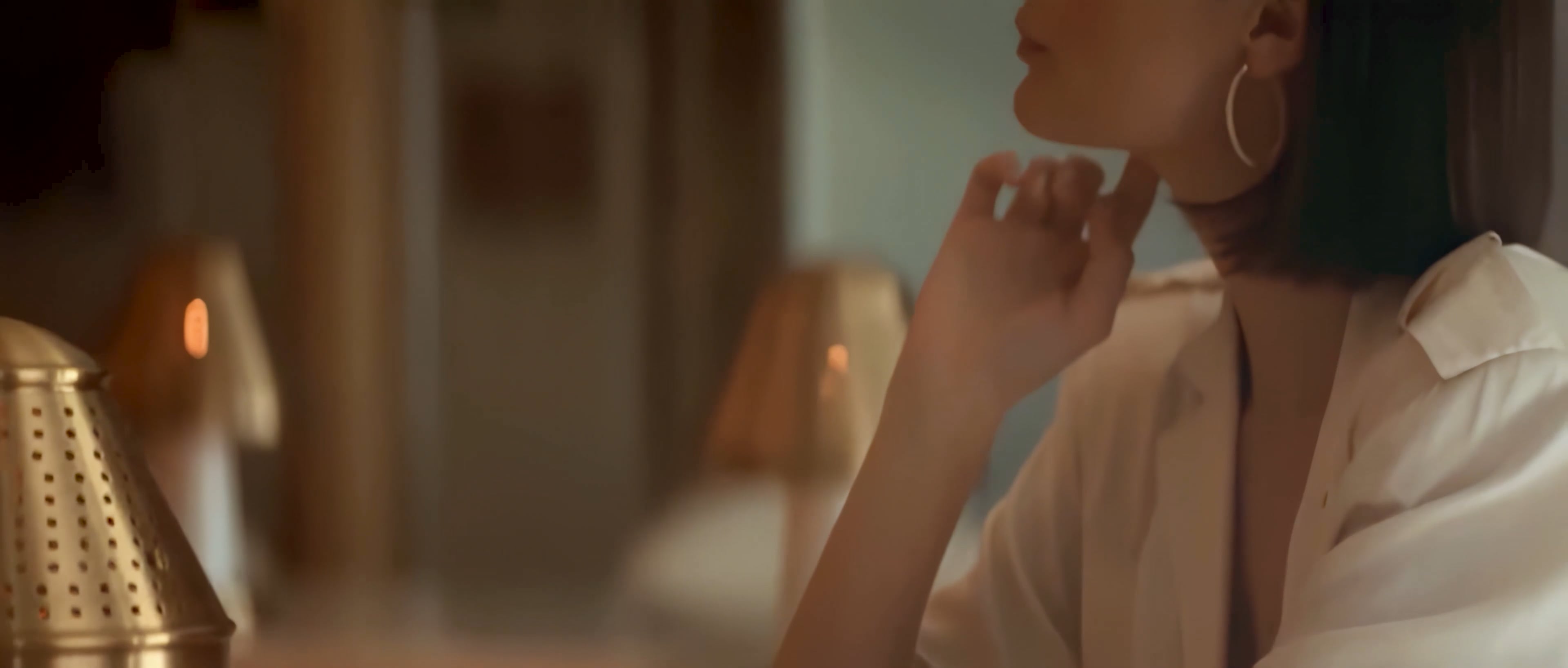 Vista laterale parziale di una donna che si tocca il collo, indossa orecchini a cerchio e una camicetta bianca al chiuso.