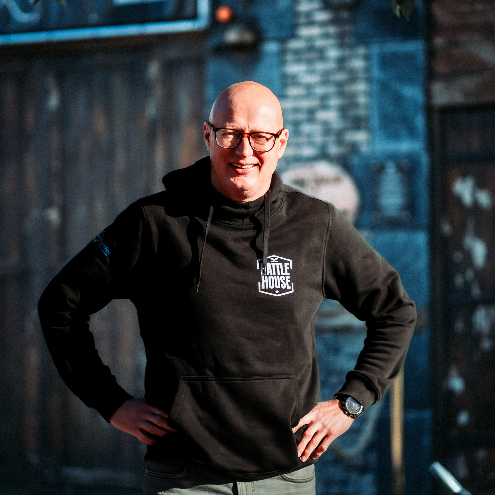 Man met een zwarte hoodie met het logo Battlehouse, glimlachend met handen in de zij voor een bakstenen muur.