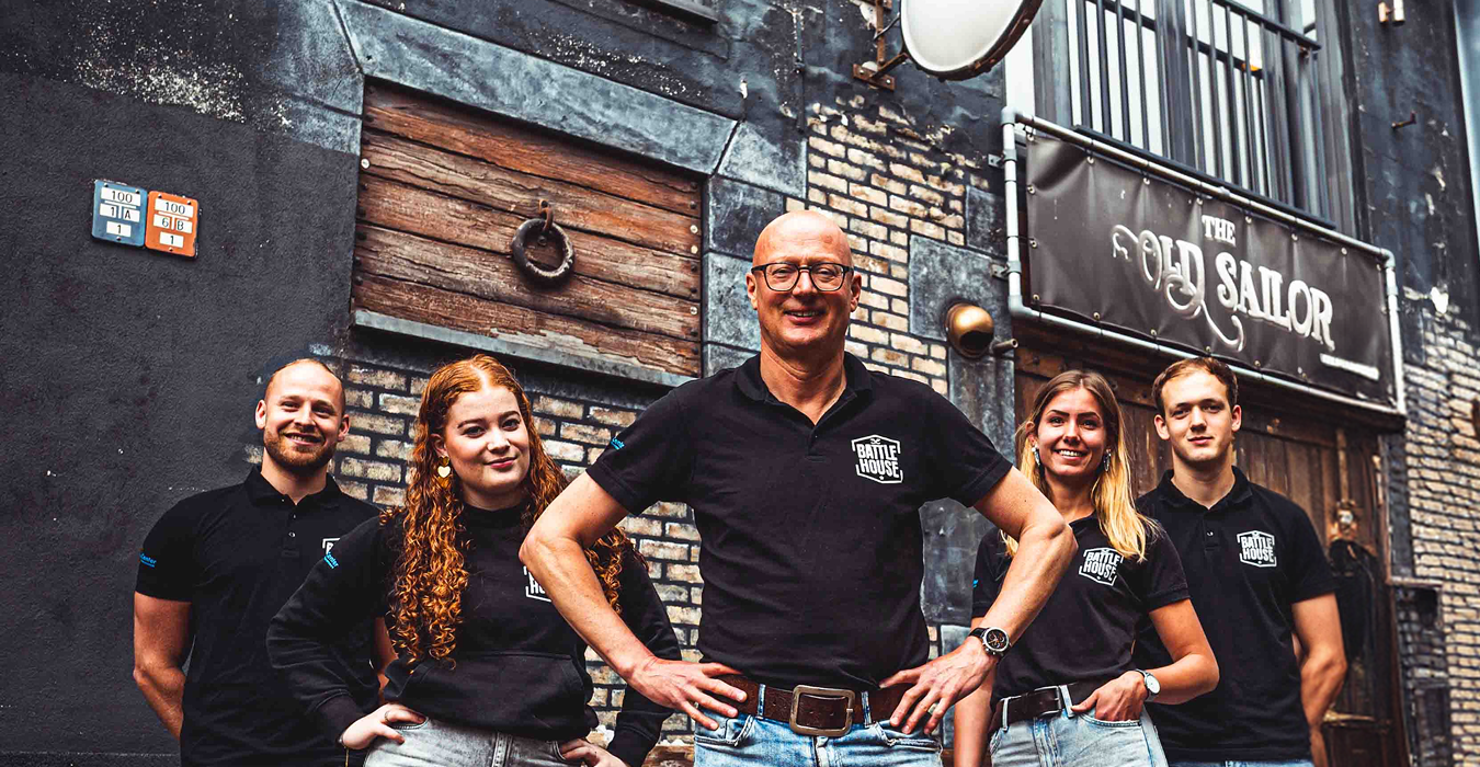 Vijf mensen in zwarte Battle House-shirts poseren lachend voor een gebouw met een bord 'The Old Sailor'.