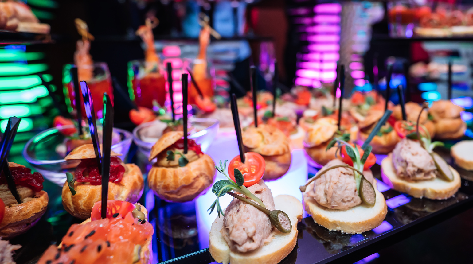 Diverse hapjes met beleg zoals zalm, mousse, tomaat en augurk op een feestelijke schaal met kleurrijke achtergrondverlichting.