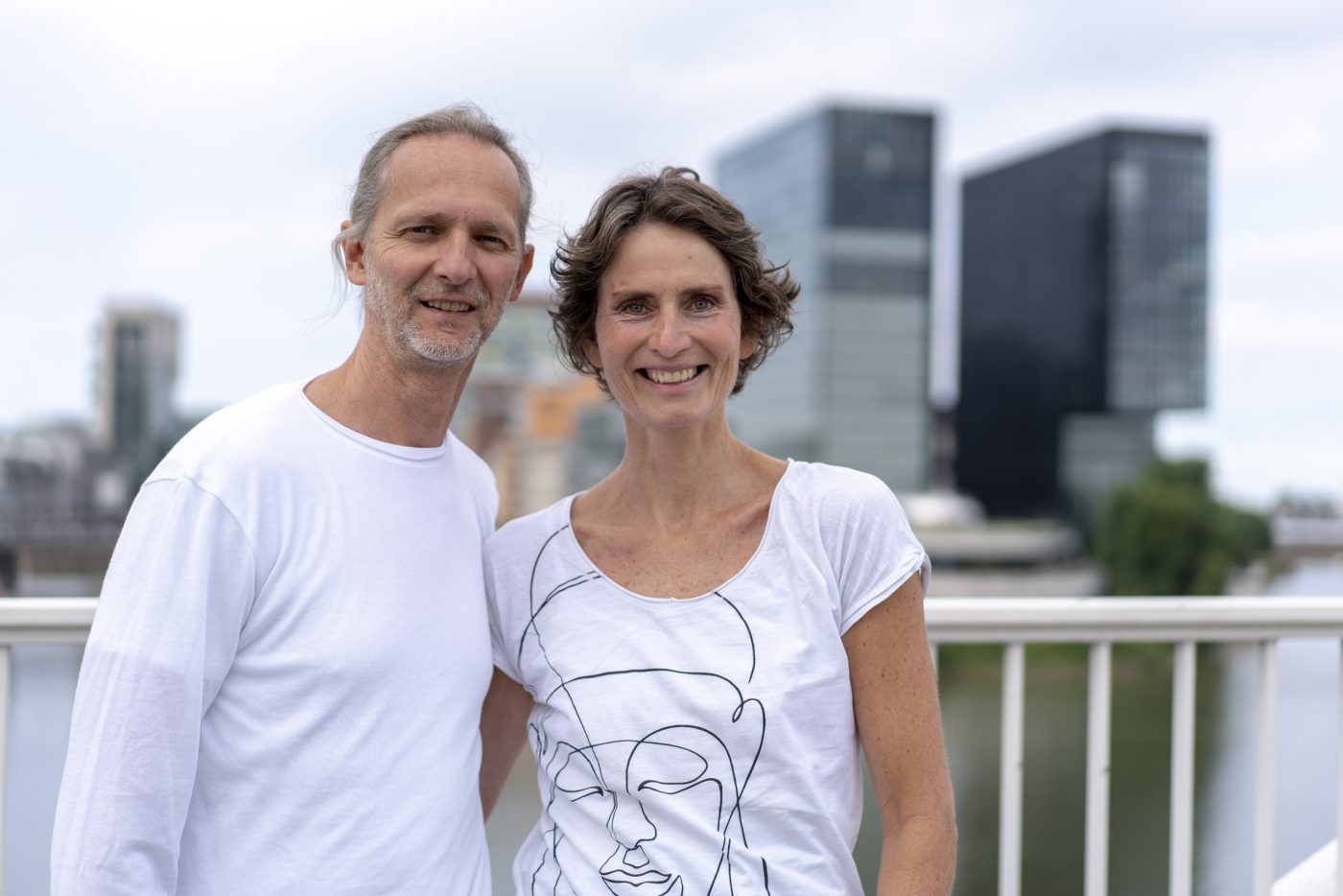 Marc und Susanne Wenke von Rundum Yoga in weißen Shirts auf einer Brücke, im Hintergrund die dunklen Hochhäuser des Medienhafens