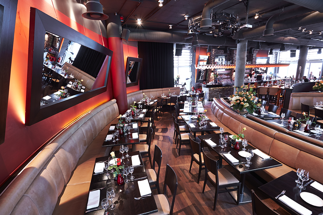 Innenraum des Riva Restaurant & Bar mit eingedeckten Tischen, geschwungenen Lederbänken und rot beleuchteter Wand