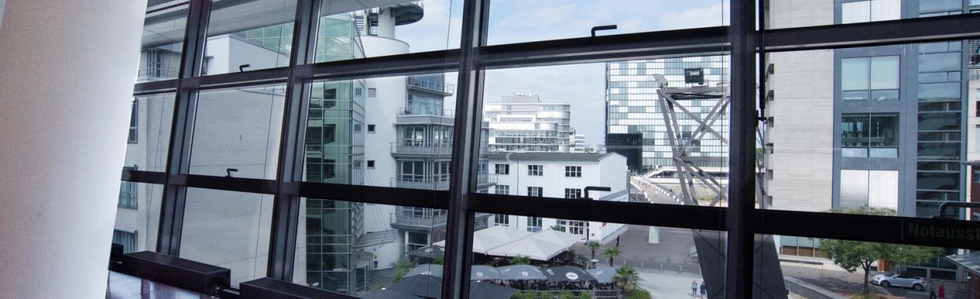 Blick durch die Fensterfront eines Büros auf die Gebäude und die Außengastronomie des Düsseldorfer Medienhafens