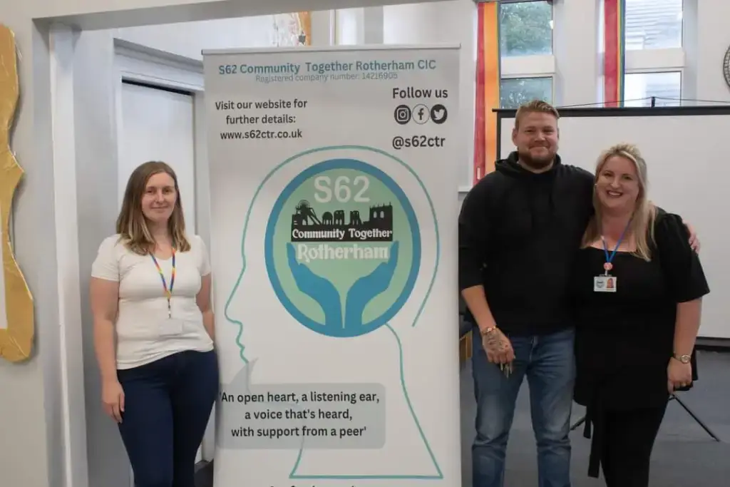 “Group of women standing outside a community venue beside a colourful ‘Women Supporting Women’ display board, smiling during an event.