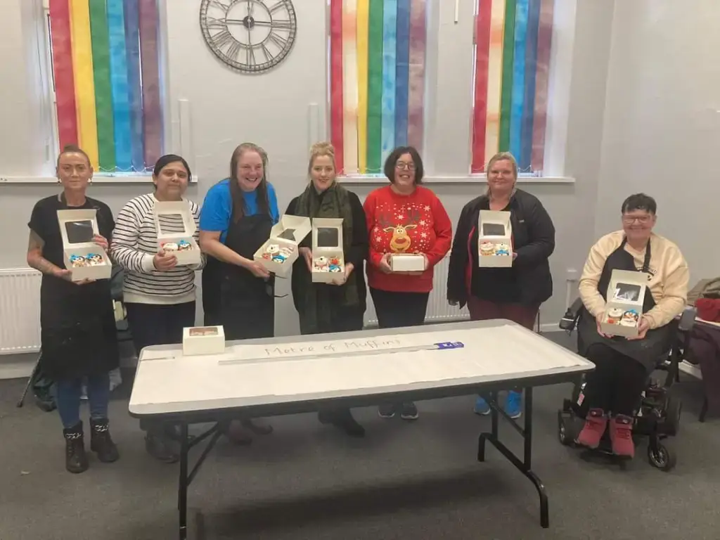 “Group of women standing outside a community venue beside a colourful ‘Women Supporting Women’ display board, smiling during an event.