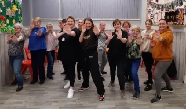 “Group of adults in a self-defence class standing in a hall, smiling and holding their hands up in a defensive stance.