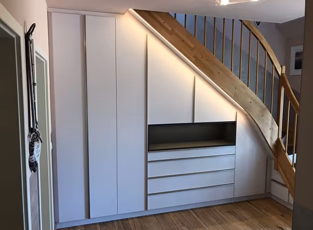 Ein maßgefertigter weißer Einbauschrank mit Schubladen und offener Ablagefläche unter einer Holzwangentreppe in einem Raum mit Holzboden.