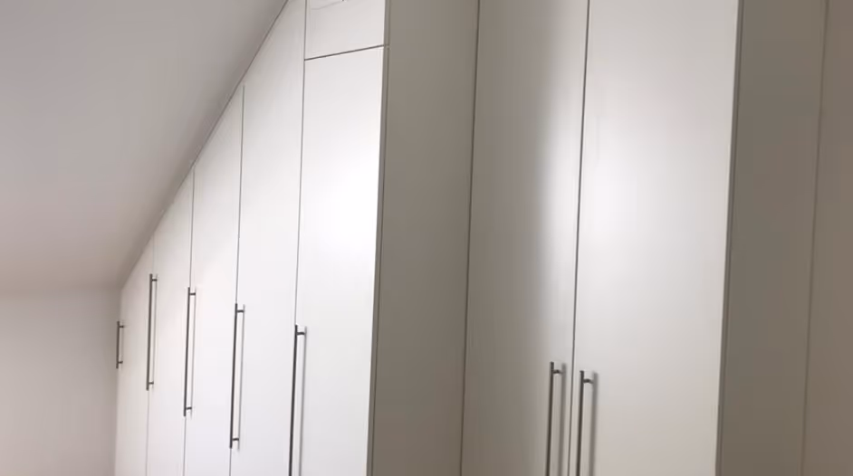 Weiße Einbauschränke mit langen Griffen an einer schrägen Wand in einem minimalistisch eingerichteten Raum.
