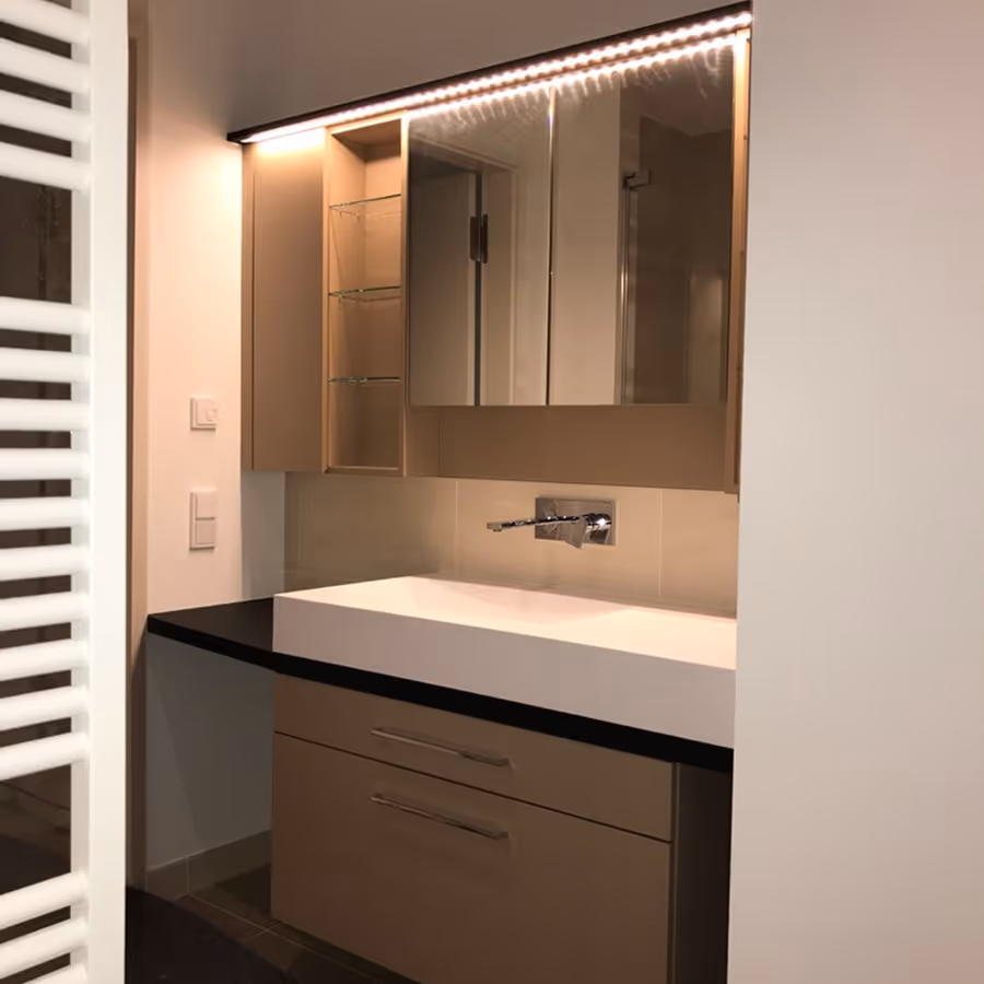 Moderner Badezimmerwaschtisch mit weißem Becken, brauner Unterschrank, beleuchtetem Spiegelschrank und Wandarmatur.