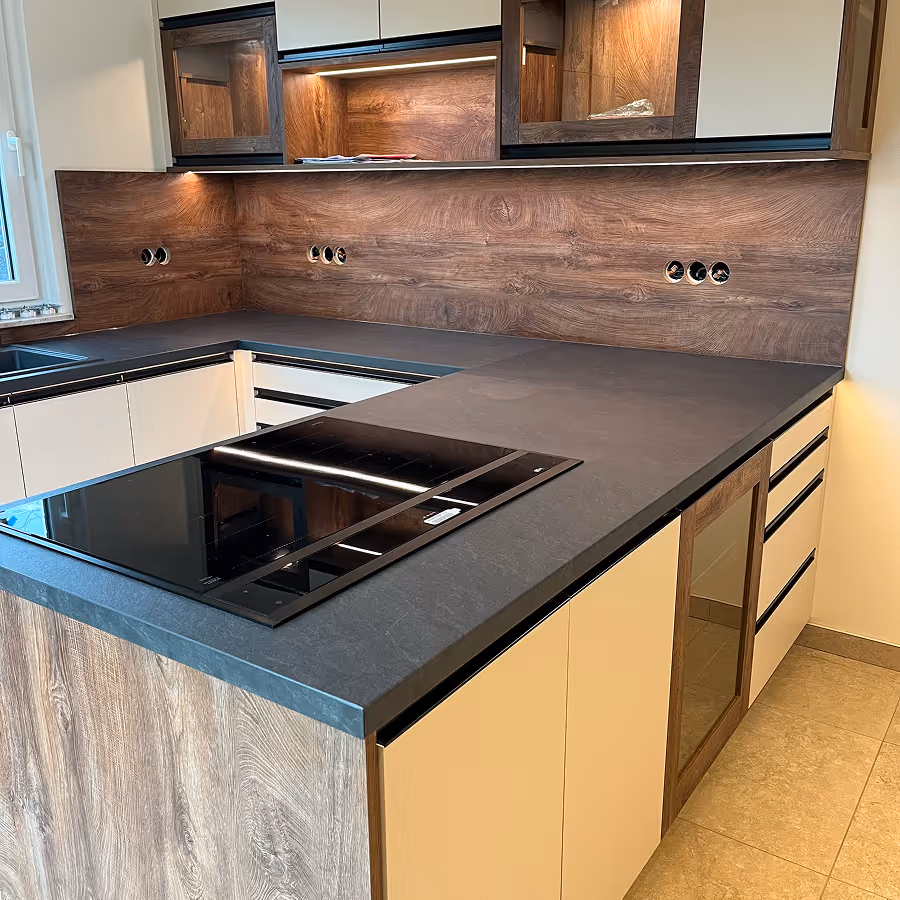 Moderne Küche mit U-förmiger Arbeitsplatte aus dunklem Material, eingebautem Kochfeld und Holzrückwand.