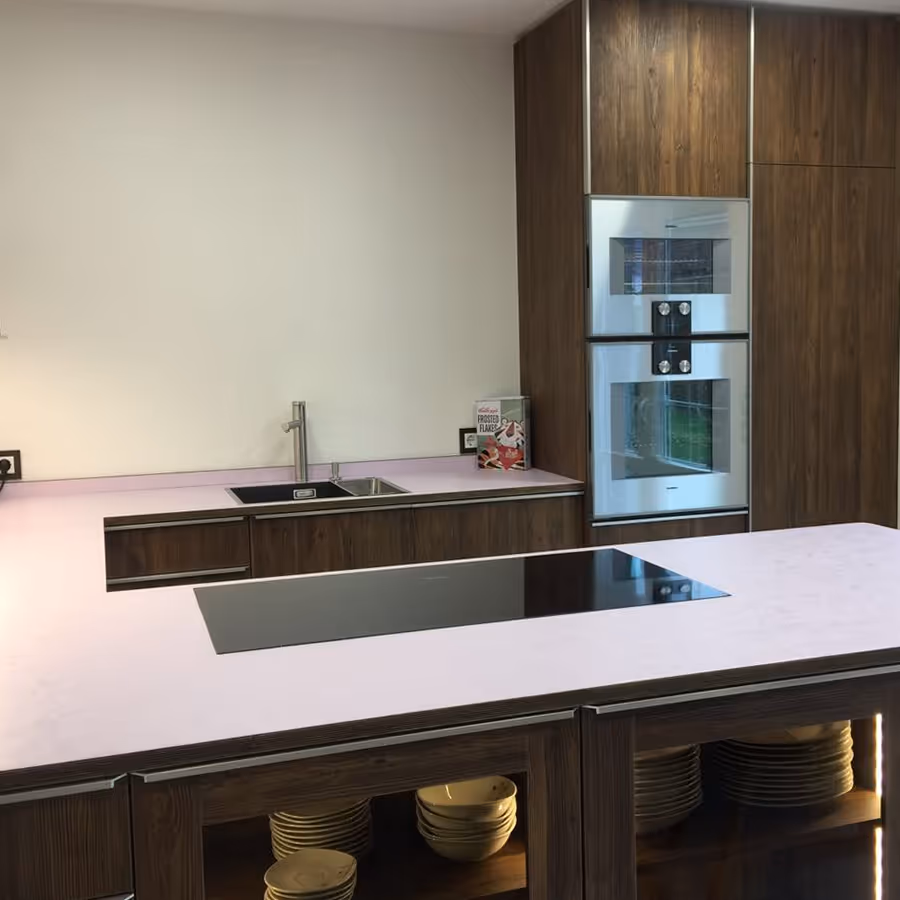 Moderne Küche mit dunklen Holzschränken, weißer Arbeitsplatte, Kochfeld integriert und Spüle mit Wasserhahn, sowie eingebauten Öfen in Holzpaneelen.
