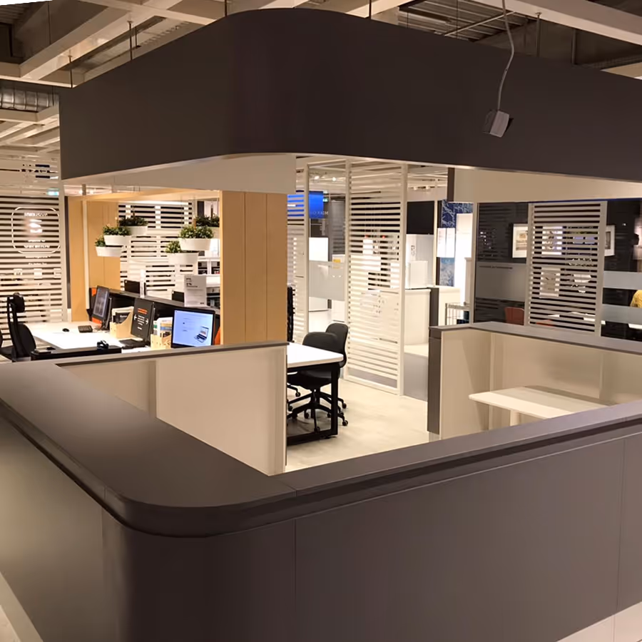 Moderne Büroecke mit mehreren Computern, schwarzen Stühlen und hängenden Pflanzen auf einem hellen Holztisch.