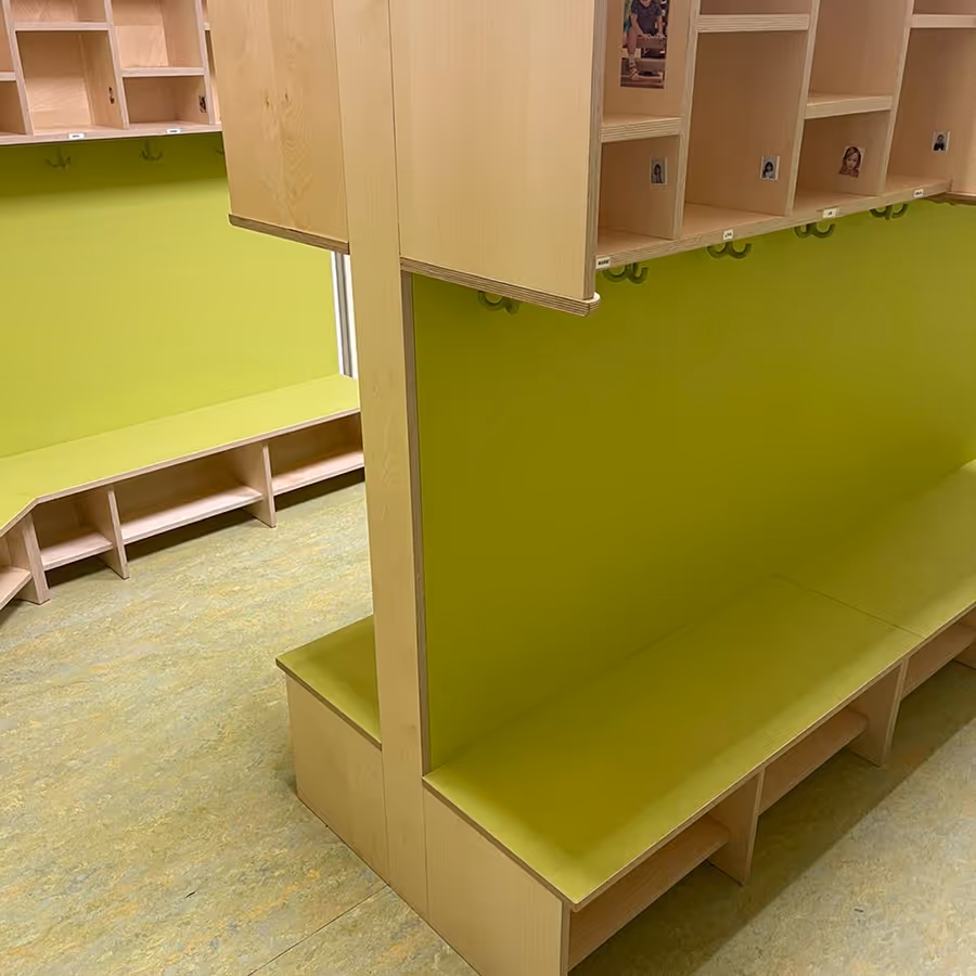 Leerer Umkleideraum mit hellgrünen Sitzbänken und Haken an der Wand, versehen mit Holzfächern darüber.
