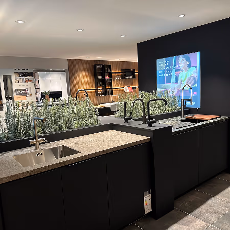 Moderne Küchenspülen mit verschiedenen Wasserhähnen in einem Showroom, umgeben von dekorativen Pflanzen.