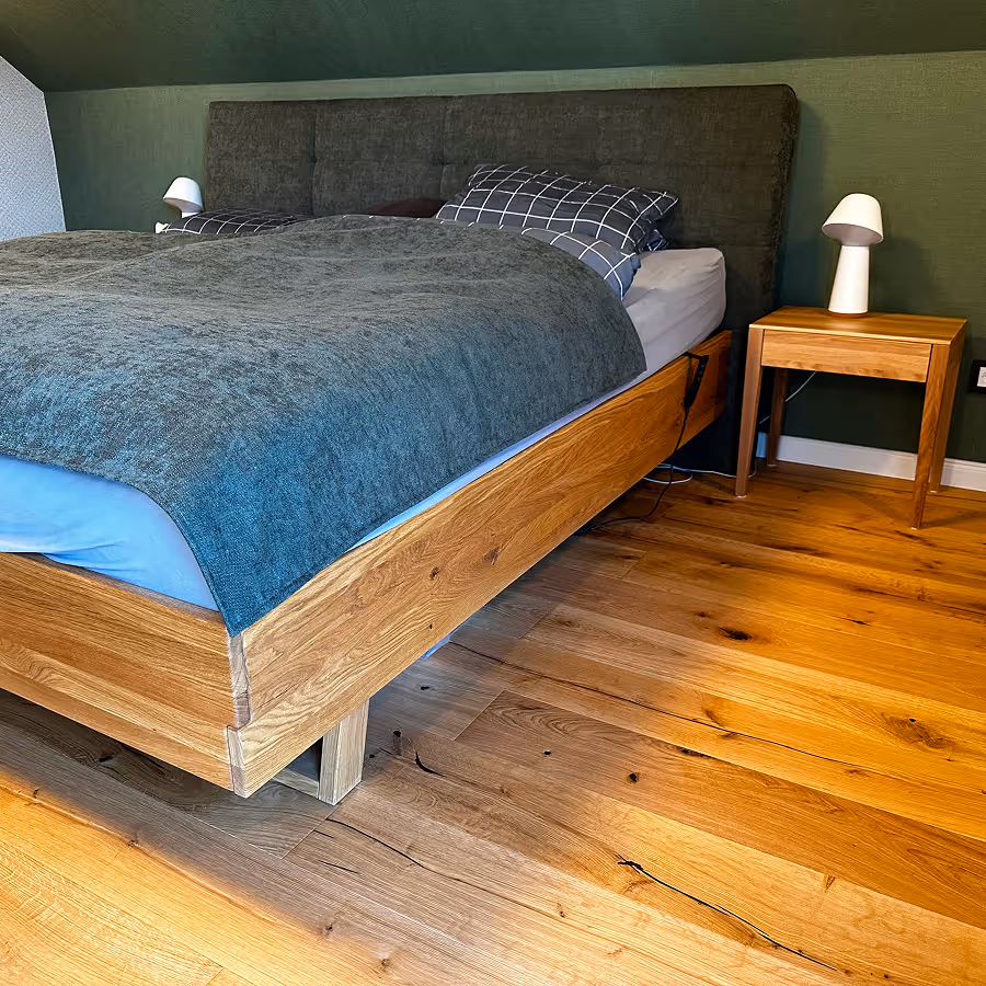 Holzbett mit dunkelgrauem Kopfteil, blauem Bettbezug und zwei karierten Kissen neben einem Holz-Nachttisch mit weißer Lampe in einem Schlafzimmer mit Holzboden.