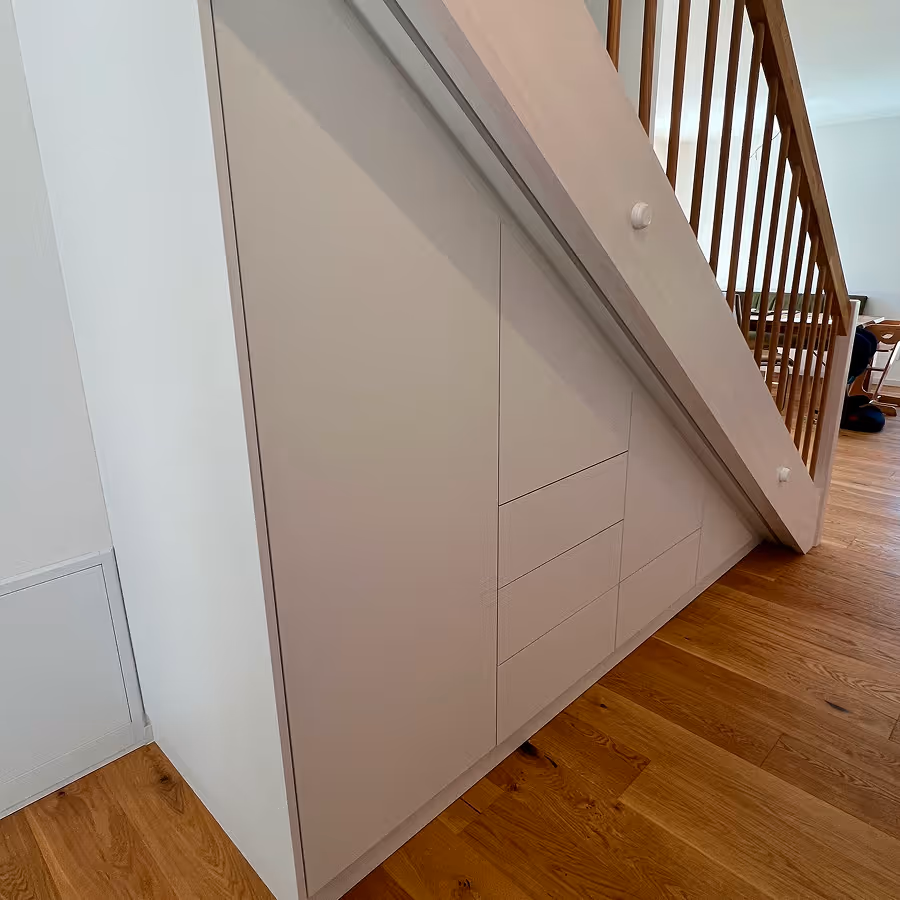 Maßgeschneiderte weiße Schranktüren und Schubladen unter einer Holztreppe mit Holzgeländer auf einem Parkettboden.