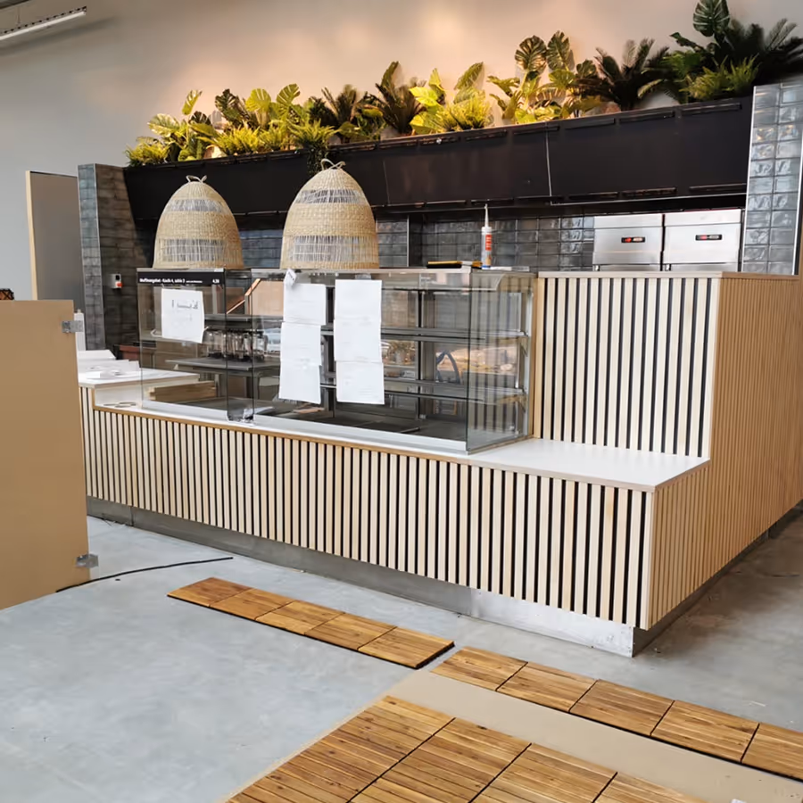 Moderne Café-Theke mit Holzverkleidung, Glasvitrine, zwei geflochtenen Hängeleuchten und grünen Pflanzen darüber.