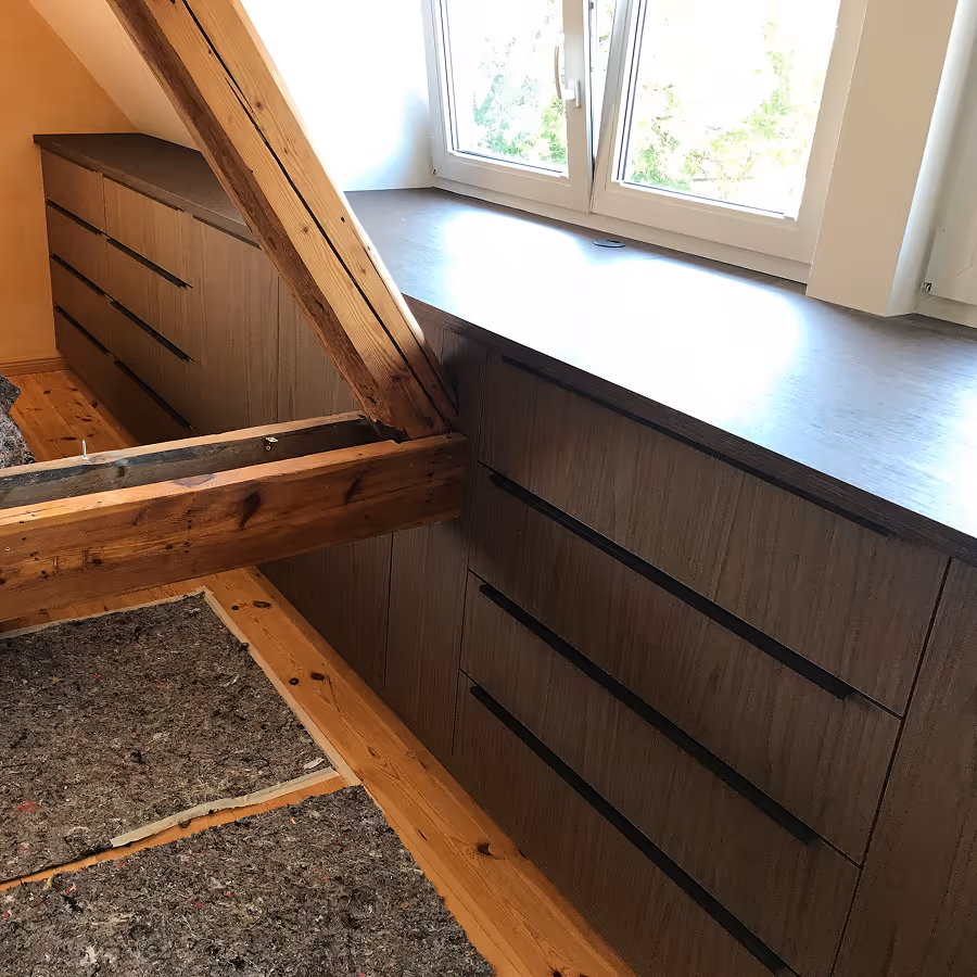 Holzkommode mit mehreren Schubladen unter einem Fenster in einem Raum mit sichtbaren Holzbalken.